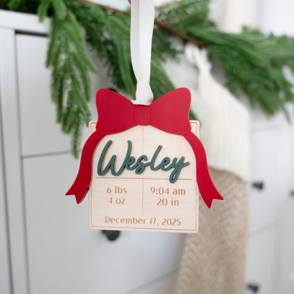 A Christmas ornament made of wood in the shape of a gift box featuring engraved baby's first name and birth stats, with a red acrylic bow