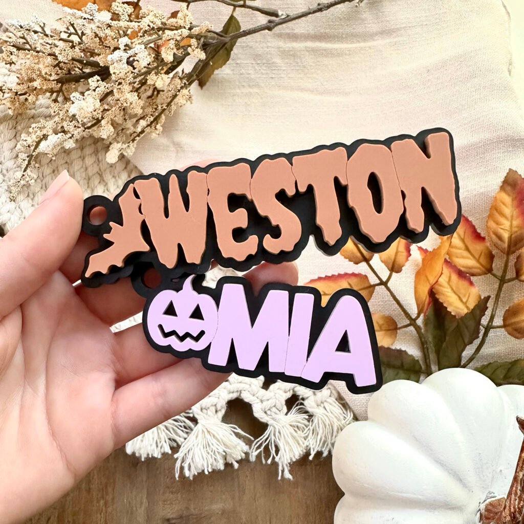 Two acrylic keychains, one in black and lavender with a pumpkin and the name Mia and the other in orange and black with a bat and the name Weston