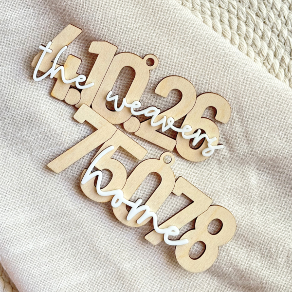 Two Christmas ornaments: A wooden Christmas ornament in the shape of the date 4.10.26 with the name the Weavers on top in white acrylic. Wooden ZIP code ornament with layered white home script across the numbers