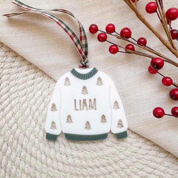 A white and green acrylic Christmas ornament in the shape of a sweater with trees and the name Liam